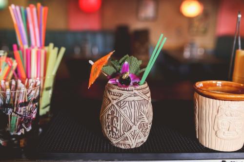 A tiki cocktail garnished with an umbrella and flowers, surrounded by colorful straws and other drinks on a bar.