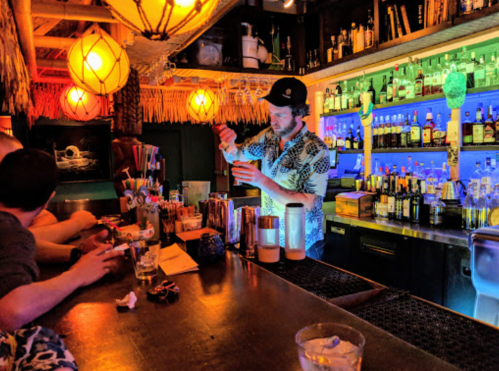 A bartender in a tropical shirt mixes drinks at a vibrant bar with colorful lighting and shelves of liquor.