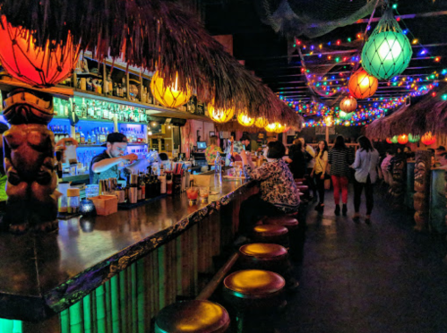 A vibrant tiki bar with colorful lights, tropical decor, and patrons enjoying drinks at the counter.