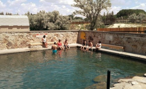 A group of people relaxes by a natural hot spring, sitting on benches around the water under a sunny sky.