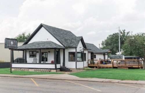 A charming white house with a dark roof, featuring a porch and a sign that reads "Firewater." Green lawn and cloudy sky.