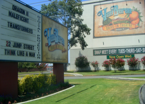 A drive-in theater sign displays movie titles and showtimes, with a large mural on the building behind it.