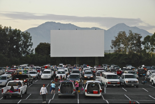 A busy drive-in movie theater with cars parked in front of a large blank screen and mountains in the background.