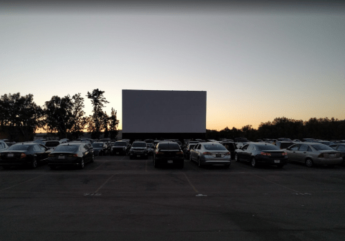 A drive-in movie theater at dusk, featuring a large blank screen and rows of parked cars.