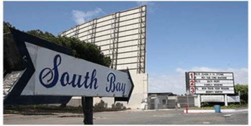 Sign for South Bay Drive-In with a large screen in the background and a movie schedule board nearby.