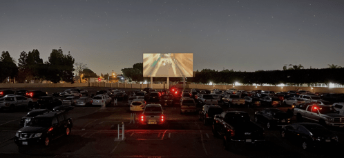 A nighttime view of a drive-in theater with cars parked and a movie screen displaying a scene under a starry sky.
