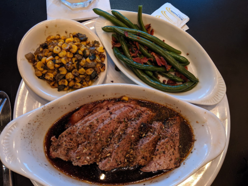 A plate with sliced beef in sauce, green beans with bacon, and a side of corn and vegetables.