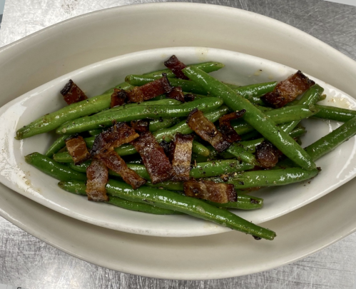 A dish of sautéed green beans topped with crispy bacon pieces, served in a white oval bowl.