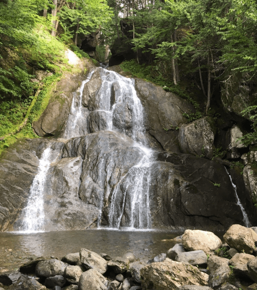 A serene waterfall cascading over rocky terrain, surrounded by lush green trees and a calm pool at the base.