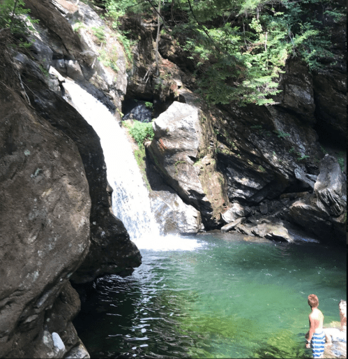 A serene waterfall cascades into a clear pool, surrounded by rocky cliffs and lush greenery, with a person standing nearby.