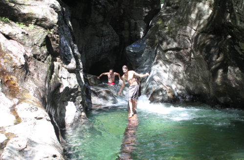 Two people navigating a rocky stream, one balancing on a log while the other wades in the water. Sunlight filters through trees.