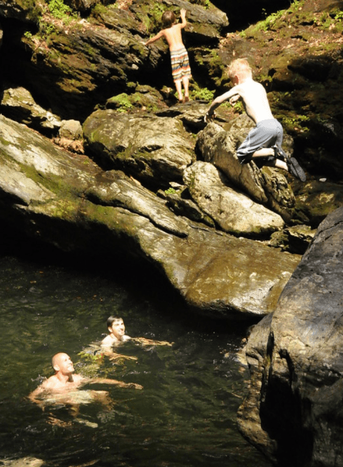 Two adults swim in a rocky pool while two children play on the rocks above, enjoying a sunny day outdoors.