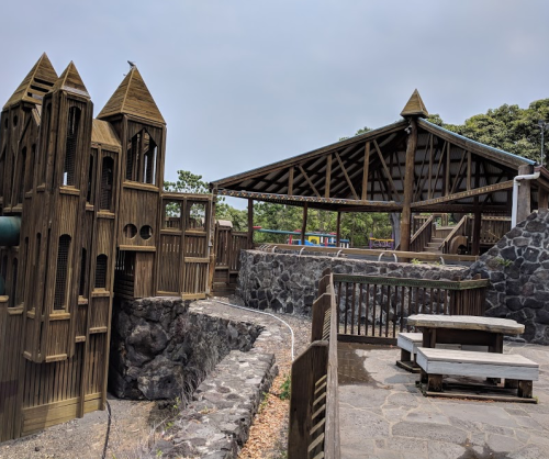 A wooden playground with a castle structure and a covered pavilion, surrounded by stone pathways and greenery.