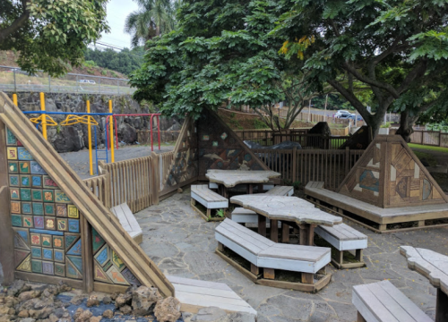 A playground area with wooden seating, colorful wall panels, and trees, surrounded by a fence and play equipment in the background.