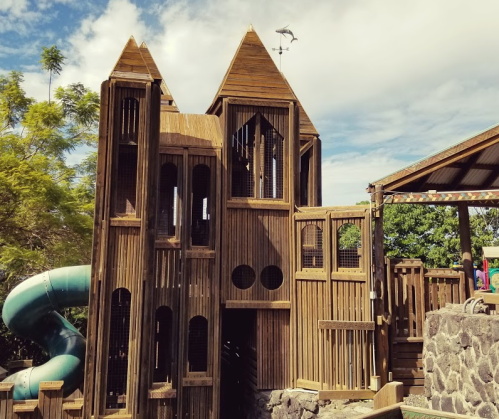 A wooden play structure resembling a castle, featuring slides and multiple levels, set against a blue sky.