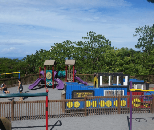 Colorful playground with slides, climbing structures, and a sandbox, surrounded by trees and a clear blue sky.