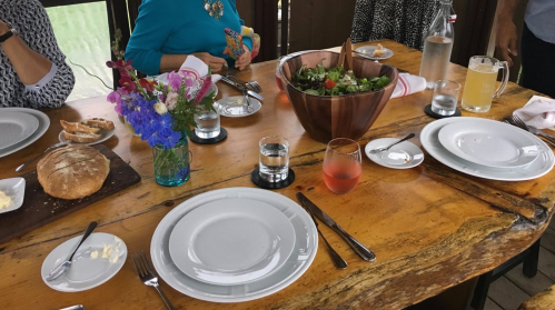 A dining table set with plates, glasses, a salad bowl, bread, and flowers, surrounded by people.
