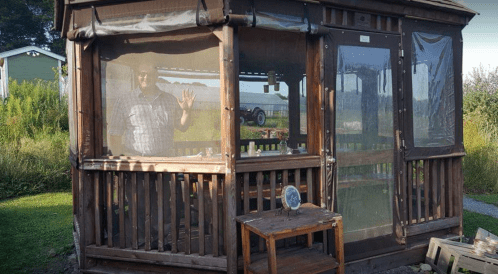 A man waves from inside a wooden gazebo with mesh screens, surrounded by greenery and a tractor in the background.