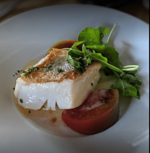 A beautifully plated piece of fish with greens and slices of tomato on a white dish.