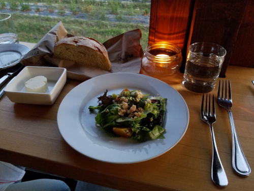 A plate of mixed greens salad with toppings, a slice of bread, butter, a glass of water, and a candle on a wooden table.