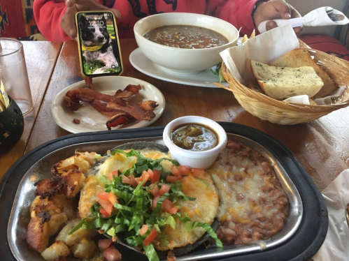 A table with a sizzling plate of eggs and potatoes, a bowl of soup, bacon, and a basket of bread.