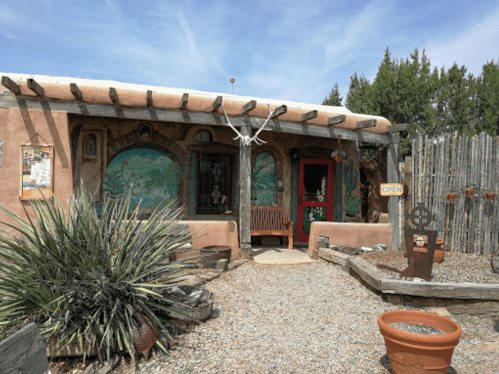 A rustic adobe building with colorful murals, a wooden bench, and a sign reading "OPEN" in a desert landscape.