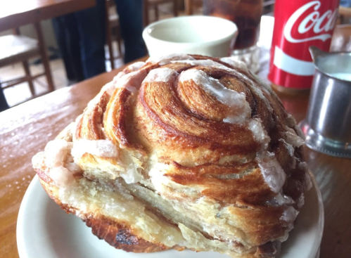 A close-up of a glazed cinnamon roll on a plate, with a cup and a soda can in the background.