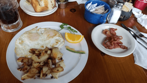 A plate with two fried eggs, seasoned potatoes, orange slice, and avocado, alongside a plate of crispy bacon.