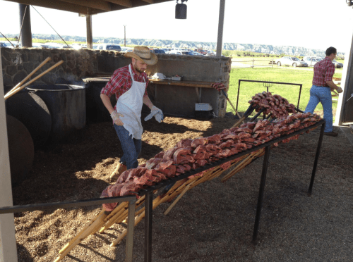A man in a straw hat and apron prepares meat on a grill outdoors, with another person in the background.