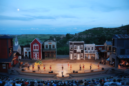A stage performance in a western-themed outdoor theater, with actors in costumes and a scenic backdrop of hills.