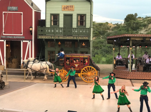A lively outdoor performance with dancers in green dresses, a horse-drawn wagon, and a band on stage in a western setting.