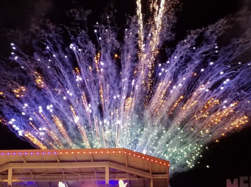 Colorful fireworks burst in the night sky above a stage, creating a vibrant display of lights and colors.