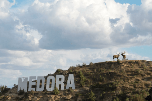 A hillside with large letters spelling "MEDORA" and two deer statues against a cloudy sky.