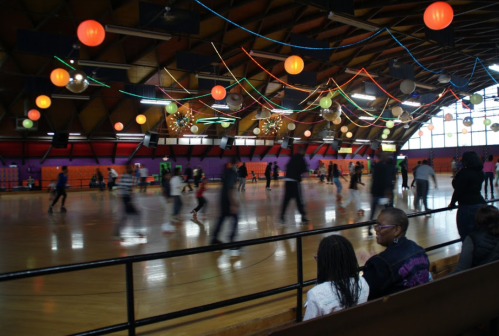 A lively roller skating rink with colorful lights and decorations, featuring skaters in motion and spectators watching.