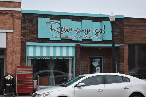 A storefront with a teal sign reading "Retro-a-go-go! Your American Pop Gift Shop Since 2004" and a parked white car.