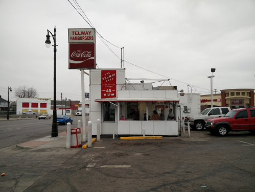 A small hamburger stand with a sign reading "Telway Hamburgers," surrounded by a parking lot and cloudy sky.