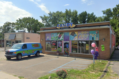 A colorful candy store with a decorated exterior and a van parked outside, surrounded by trees and a clear sky.