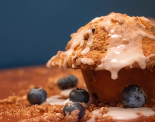A close-up of a blueberry muffin drizzled with icing, surrounded by fresh blueberries and crumbs on a wooden surface.