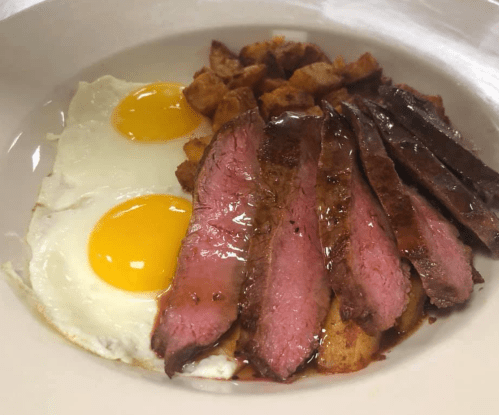 A plate of breakfast featuring two sunny-side-up eggs, crispy potatoes, and slices of medium-rare steak.