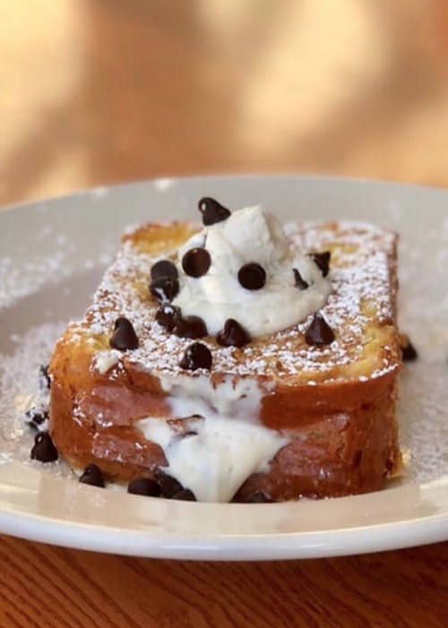 A slice of French toast topped with whipped cream and chocolate chips, dusted with powdered sugar on a white plate.