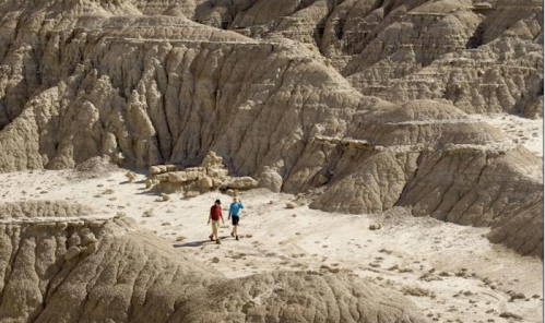 Two hikers explore rugged, dry terrain with layered rock formations under a clear blue sky.
