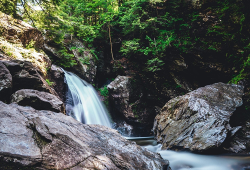 A serene waterfall cascades over rocks, surrounded by lush green trees in a tranquil forest setting.