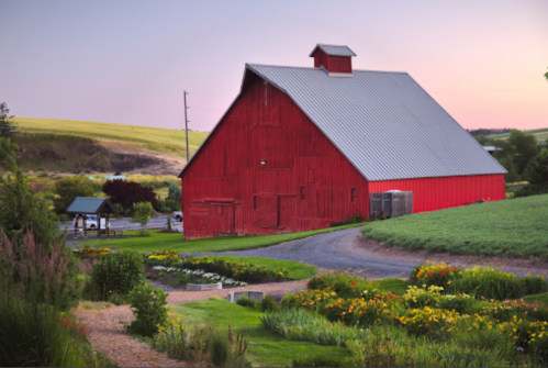 A vibrant red barn stands beside a winding path and colorful flower beds, with rolling hills in the background at sunset.