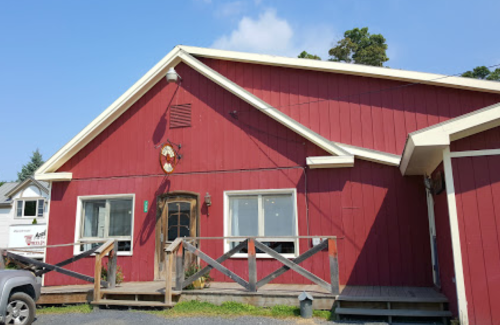 A red wooden building with a sloped roof, featuring large windows and a wooden railing in front.