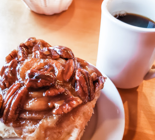 A sticky pecan roll on a plate next to a white cup of coffee on a wooden table.