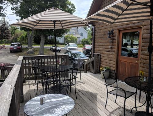 A wooden deck with striped umbrellas, tables, and chairs, overlooking parked cars and greenery.