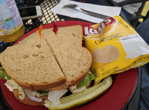 A sandwich with lettuce, tomato, and onion on whole grain bread, served with a side of Lay's classic potato chips and a pickle.
