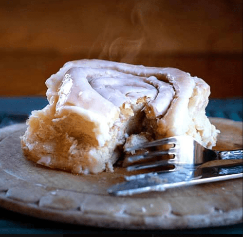 A freshly baked cinnamon roll with icing, partially cut, resting on a wooden board with steam rising.