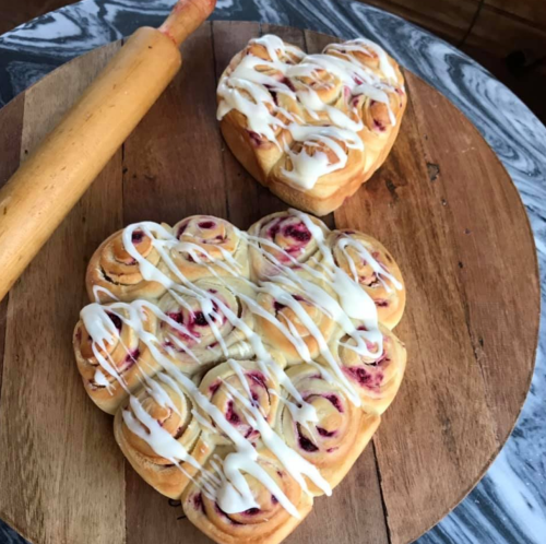 Two heart-shaped cinnamon rolls drizzled with icing, placed on a wooden cutting board next to a rolling pin.