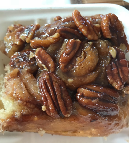 A close-up of a sticky pecan roll topped with caramel and whole pecans on a white plate.